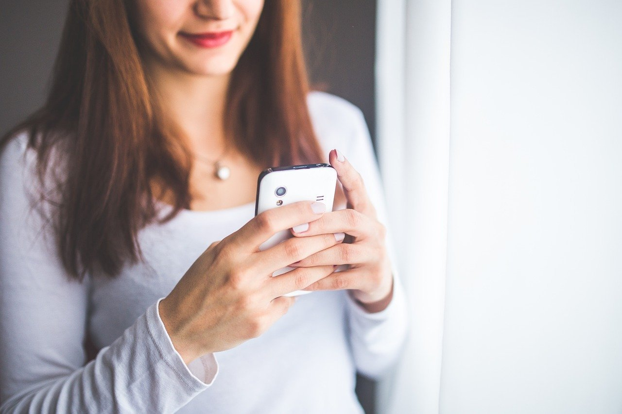 Beautiful young woman with straight brunette hair wearing white long sleeve shirt holding white cell phone