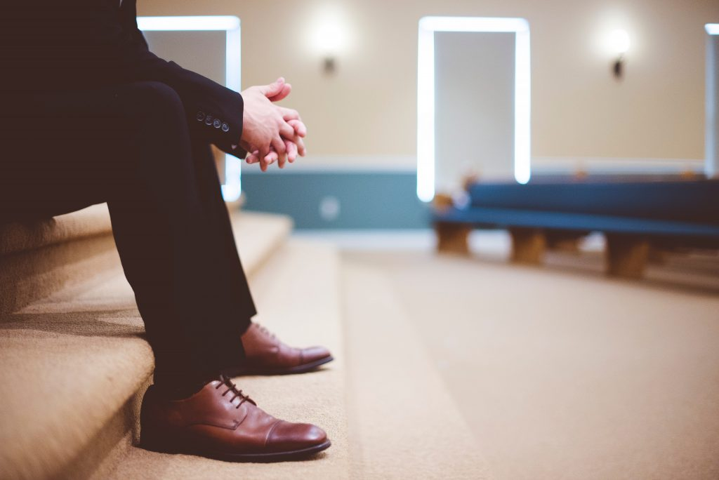 Closeup of man sitting on steps in church wearing black dress slacks and brown leather shoes with hands folded in prayer