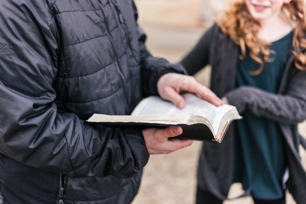 Close up of man in a dark grey winter jacket holding an open Bible with a smiling young girl with red curls and dark grey and green jacket.